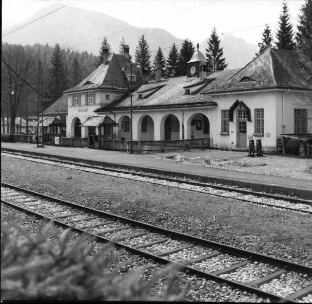 Ein Schwarz-Weiß-Bild zeigt einen alten Bahnhof mit einem Glockenturm, umgeben von Bäumen und Bergen im Hintergrund.