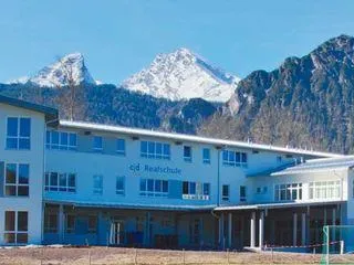 Ein großes, modernes weißes Gebäude mit der Aufschrift 'cd Realschule' und vielen Fenstern befindet sich am Fuße verschneiter Berge.