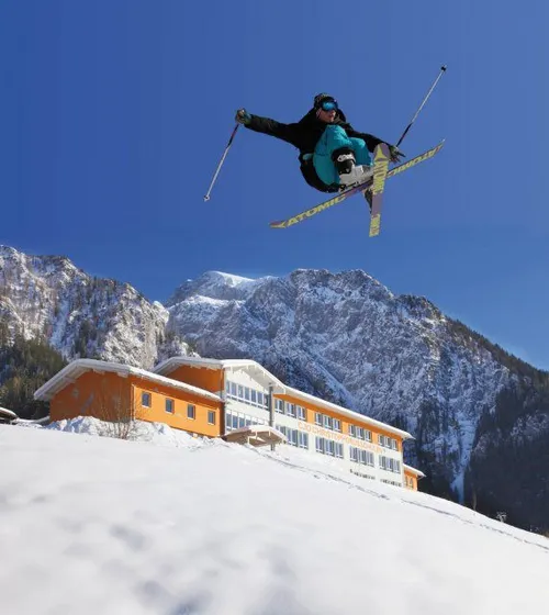 Ein Skifahrer springt in der Luft, hält Skistöcke in der Hand, trägt eine blaue Jacke und eine Schutzbrille, mit einem verschneiten Berg im Hintergrund.