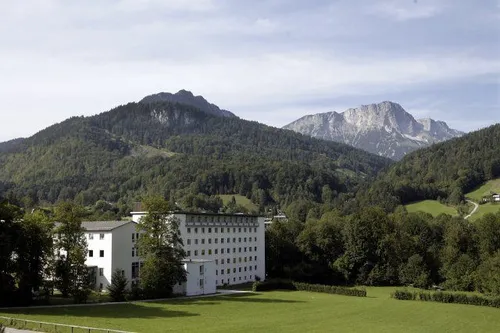 Ein großes weißes Gebäude steht auf einem Hügel, umgeben von einem üppigen grünen Feld und Bäumen. Im Hintergrund befinden sich Berge mit felsigen Klippen und dichten Wäldern.
