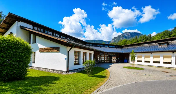Ein Gebäude mit vielen Fenstern steht unter einem teilweise bewölkten Himmel. Es hat eine gepflasterte Auffahrt, die zu ihm führt und ist von einem grünen Rasen umgeben.