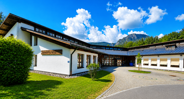 Ein Gebäude mit vielen Fenstern steht unter einem teilweise bewölkten Himmel. Es hat eine gepflasterte Auffahrt, die zu ihm führt und ist von einem grünen Rasen umgeben.