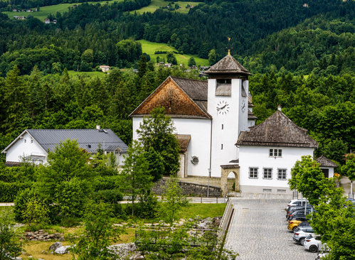 Eine weiße Kirche mit einem braunen Dach steht in einem grünen Tal, umgeben von Bäumen. Sie verfügt über einen Glockenturm und einen Uhrturm, mit geparkten Autos in der Nähe.