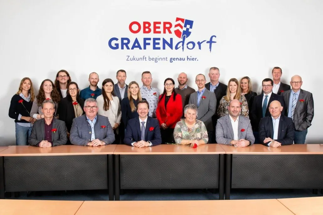 Eine Gruppenaufnahme von Menschen in formeller Kleidung mit einer roten Rose, die an ihren Anzügen befestigt ist, stehen vor einem Schreibtisch und einem Banner mit dem Text 'OBER GRAFENDORF Zukunft beginnt genau hier.'