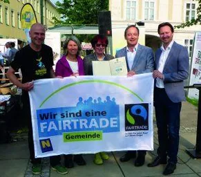 Fünf Personen stehen zusammen und halten ein Banner, auf dem steht 'Wir sind eine Fairtrade-Gemeinde' auf Deutsch. Das Banner zeigt das Fairtrade-Logo und sie sind draußen in der Nähe eines Gebäudes.