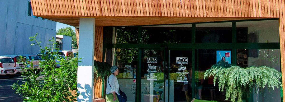 Ein Geschäft mit Holzpaneelen und Glastüren ist zu sehen. Eine Frau geht daran vorbei. Auf den Glastüren befinden sich Schilder, die 'Styx Welcome Center' und 'Styx Shop' anzeigen.