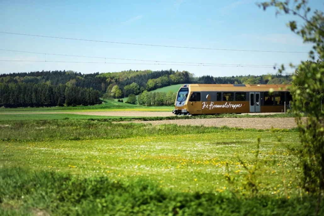 Ein goldener Zug namens 'Heimwehstrappen' fährt auf einer Strecke durch eine üppige grüne Landschaft mit Bäumen und einem klaren blauen Himmel.