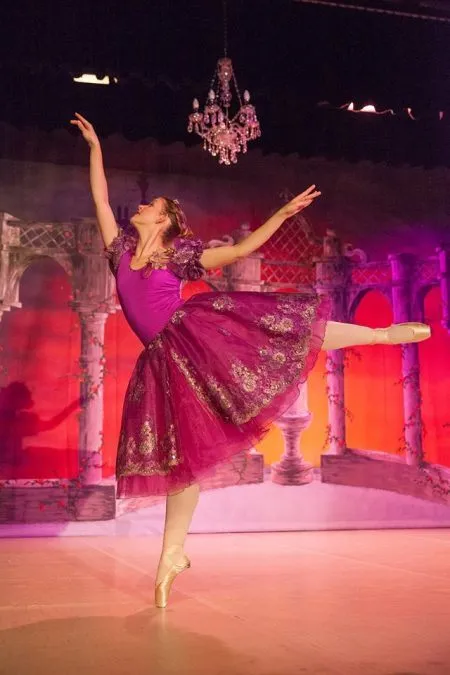 Eine Balletttänzerin in einem lila Tutu führt auf einer Bühne mit einem Hintergrund aus roter und lila Wand auf.