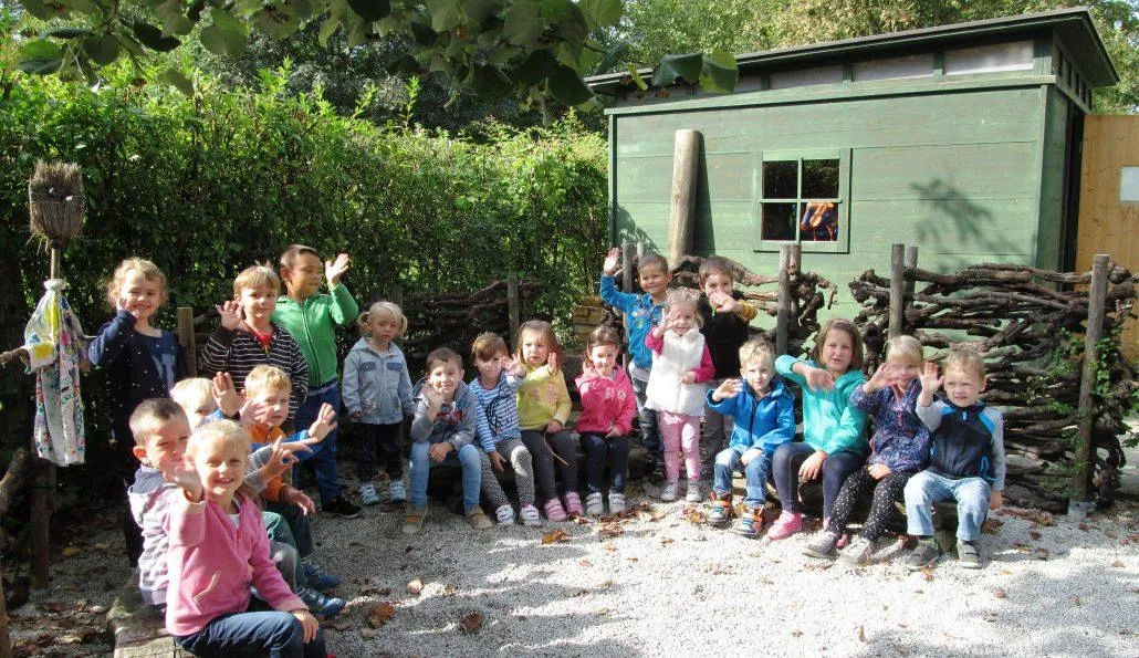 Eine Gruppe von Kindern sitzt lächelnd und winkend im Freien auf dem Boden, mit einem grünen Schuppen und Pflanzen im Hintergrund.