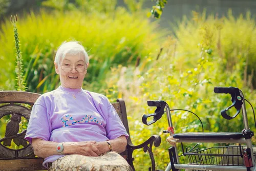 Eine ältere Frau mit Brille und einem lila T-Shirt mit dem Wort Bella darauf sitzt auf einer Bank in einem Garten mit einem Fahrrad neben ihr.