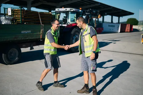 Zwei Männer in Warnwesten schütteln sich die Hände in einem Logistikbereich mit einem Traktor und einem Lkw im Hintergrund.