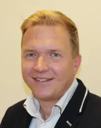 Ein Mann mit blonden Haaren, blauen Augen und einem freundlichen Lächeln. Er trägt ein weißes Hemd und einen schwarzen Blazer.