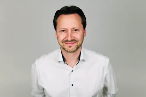 Ein Mann mit kurzen dunklen Haaren und einem Bart lächelt und posiert für ein Foto. Er trägt ein weißes Hemd.