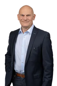 Ein Mann in einem dunkelblauen Anzug und hellblauem Hemd steht vor schwarzem Hintergrund und lächelt.