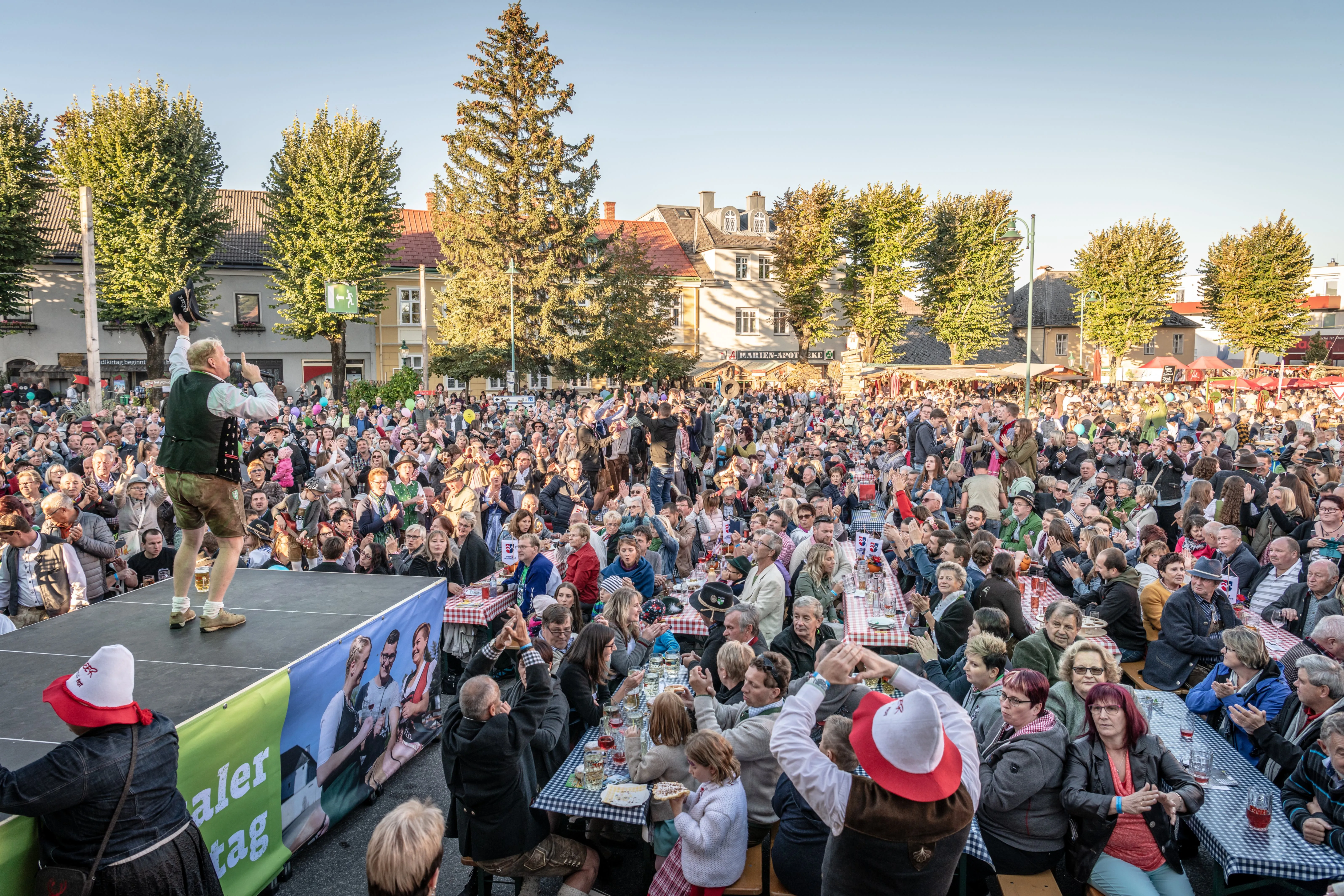 Eine große Menschenmenge versammelt sich im Freien für eine Veranstaltung, mit Leuten an Tischen, stehend und jubelnd. Bäume und Gebäude sind im Hintergrund zu sehen.