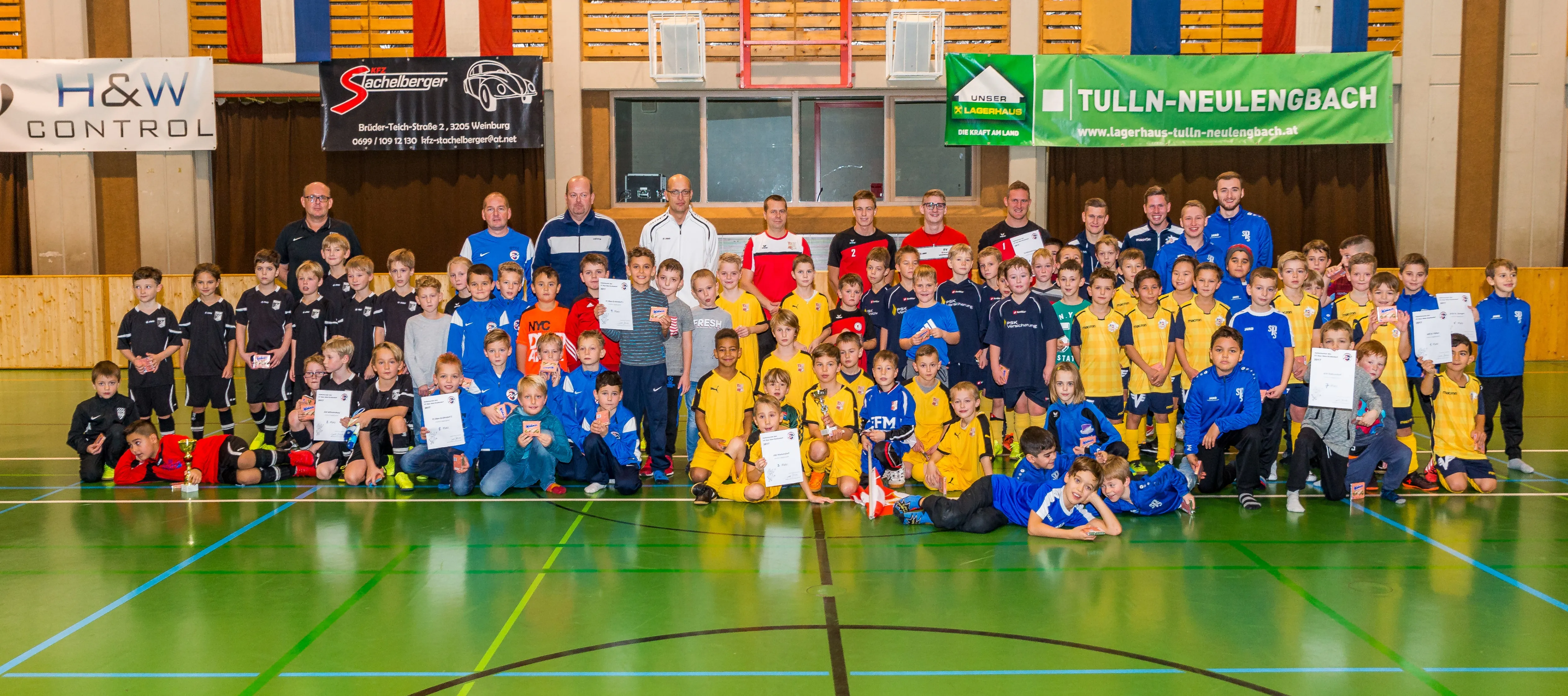 Gruppe junger Fußballspieler, Erwachsene und Trainer posieren für ein Foto auf einem grünen Spielfeld mit Bannern. Spieler halten Urkunden und Pokale.