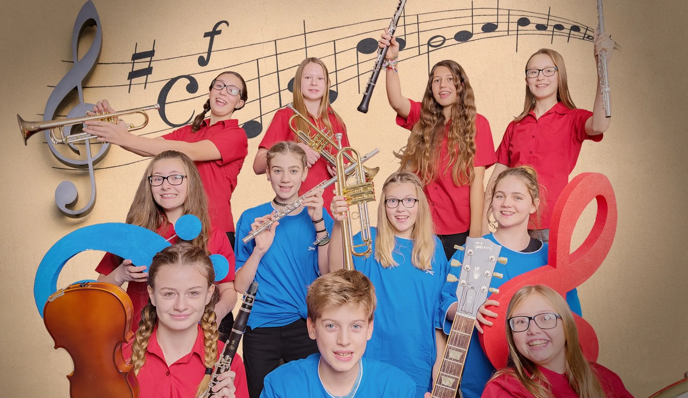Eine Gruppe junger Schüler, gekleidet in blauen und roten Hemden, halten verschiedene Musikinstrumente wie Trompeten, Flöten und eine Gitarre in der Hand und posieren für ein Foto vor einem musikalischen Notenhintergrund.