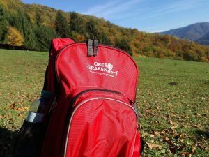 Ein roter Rucksack mit der Aufschrift 'Ober Grafentor' liegt auf dem Gras, mit Bäumen und Bergen im Hintergrund.