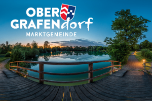 Ein szenisches Bild von Ober Grafendorf zeigt eine Holzbrücke über einem ruhigen See, einen Sonnenuntergang und einen Weg, der zu einem Baum führt.