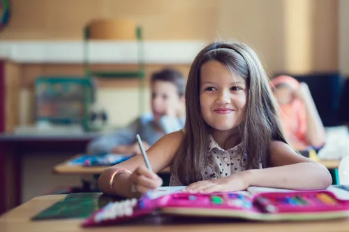 Ein kleines Mädchen schreibt in einem Klassenzimmer auf Papier mit einem Stift. Sie lächelt und andere Schüler sind im Hintergrund.