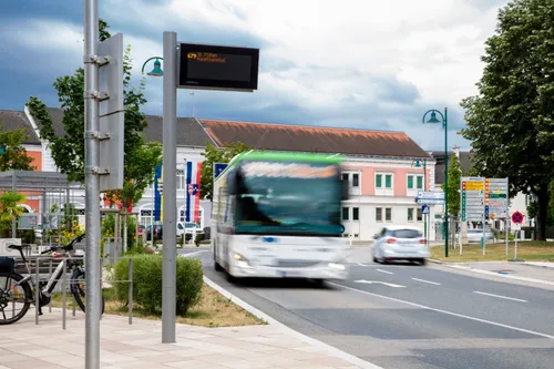 Ein Bus auf einer Straße zeigt ein digitales Display mit dem Ziel St. Pölten Hauptbahnhof. Hinter dem Bus fährt ein Auto auf der Straße. Gebäude und Bäume befinden sich an den Seiten der Straße.