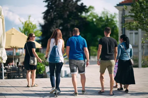 Eine Gruppe von Menschen geht an einem sonnigen Tag, zwei halten sich an den Händen und einer hat ein Einrad. Sie sind lässig gekleidet, einige tragen Sonnenbrillen. Bäume und ein Gebäude sind im Hintergrund.
