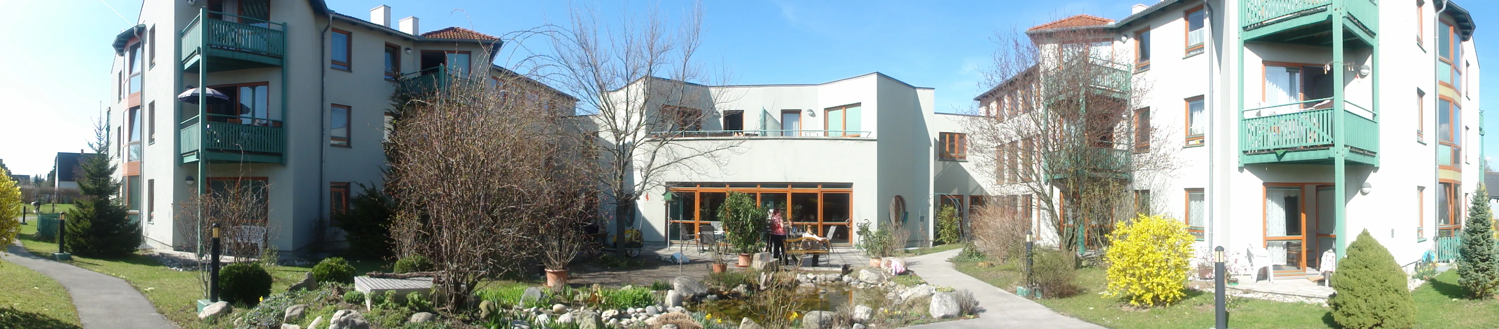 Ein modernes weißes Haus mit Balkon und Garten davor. Menschen sitzen auf einer Bank vor dem Haus.