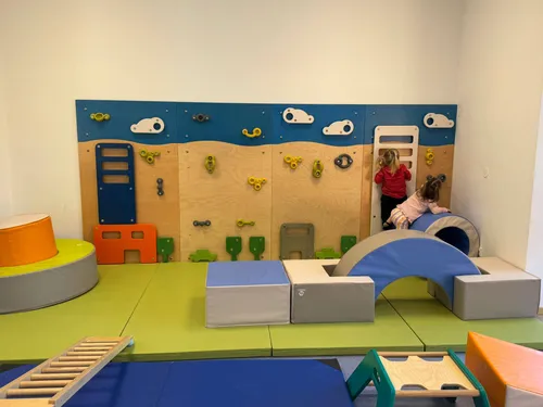Zwei Kinder spielen in einem Spielzimmer mit einer Wand voller Kletterspielzeug. Es gibt bunte Blöcke und eine blaue Rutsche.