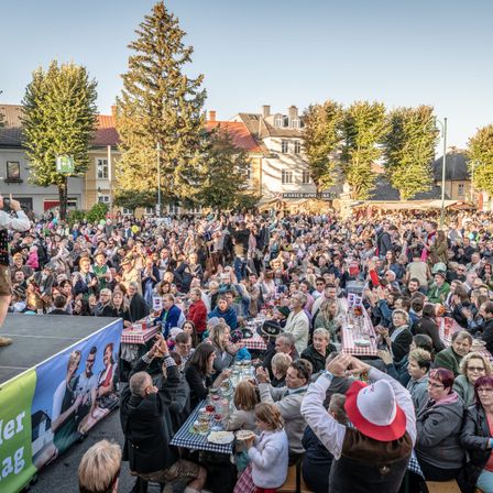 Eine große Menschenmenge versammelt sich im Freien, wahrscheinlich bei einem Festival, mit Menschen, die an Tischen sitzen, stehen und einige klatschen. Es gibt eine Bühne mit einem Banner auf der linken Seite und Bäumen und Gebäuden im Hintergrund.