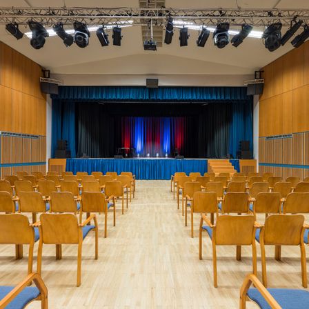 Ein leerer Saal mit blauen und roten Vorhängen, Holzstühlen und Bühnenequipment. Die Bühne hat einen blauen Hintergrund mit roten und blauen Vorhängen.