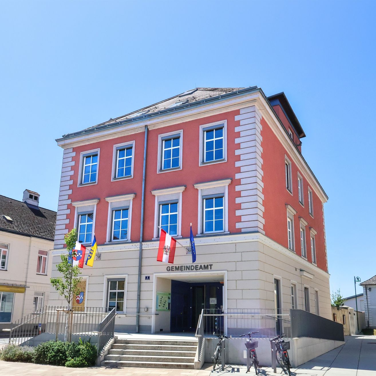Ein rotes und weißes Gebäude mit vielen Fenstern und Fahnen davor. Es hat Stufen, die zu einer Tür führen, und Fahrräder sind in der Nähe geparkt.