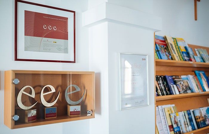 Ein Raum mit einer weißen Wand, auf der ein Holzregal mit drei Buchstaben 'e' steht. Ein gerahmtes Zertifikat ist über dem Regal montiert. Ein Holzregal voller Bücher befindet sich rechts.