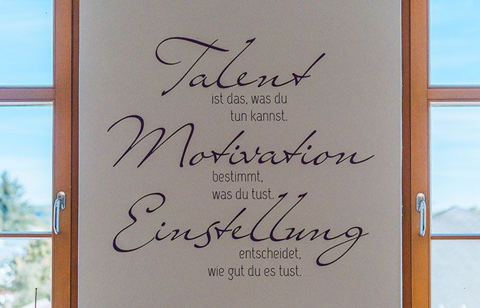 Eine weiße Wand mit einem violetten Text, der besagt: 'Talent ist, was du tun kannst, Motivation bestimmt, was du tust, Einstellung entscheidet, wie gut du es tust.' Hinter der Wand befinden sich zwei Fenster.