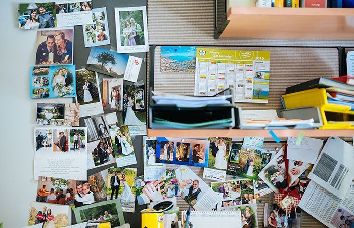 Ein Anschlagbrett ist mit Hochzeitsfotos, Notizen und einem Kalender für 2016 bedeckt. Das Brett befindet sich in der Nähe eines Schreibtischs mit Büchern und einem gelben Behälter.