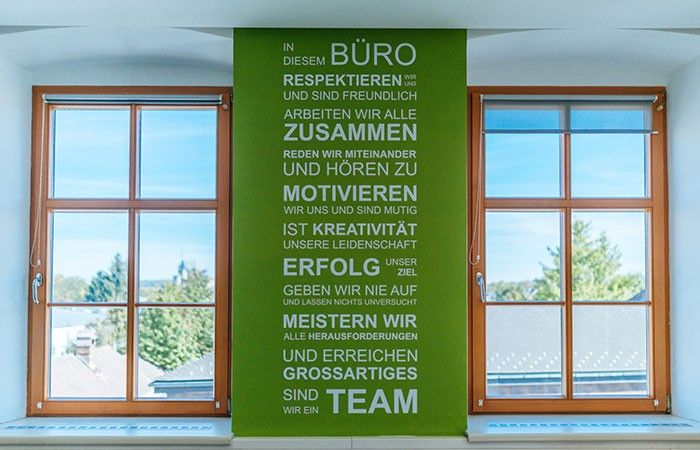 Ein grünes vertikales Schild mit weißem Text ist zwischen zwei Fenstern an einer weißen Wand befestigt. Es betont Teamarbeit und Kreativität.