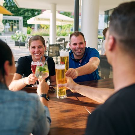 Eine Gruppe von Menschen sitzt an einem Tisch in einem Außenrestaurant. Sie clinken mit ihren Biergläsern zusammen und lächeln.