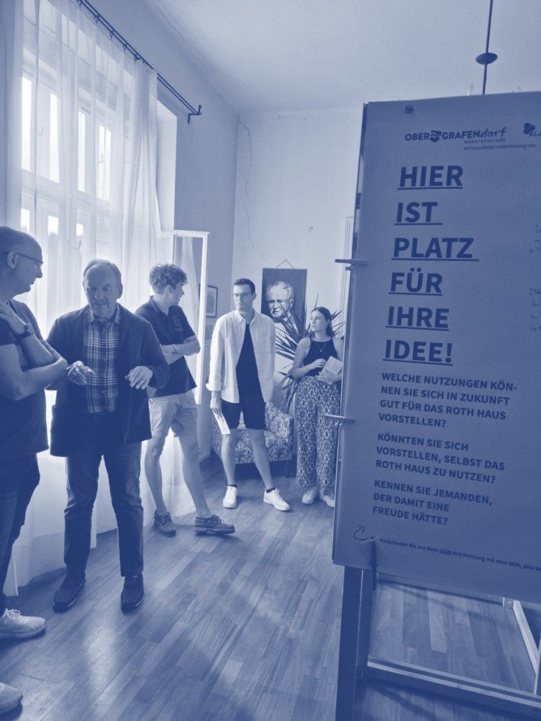 Eine Gruppe von Menschen diskutiert in der Nähe eines Schildes, das 'Hier ist der Platz für Ihre Idee!' steht. Der Raum hat einen Holzboden und weiße Wände.