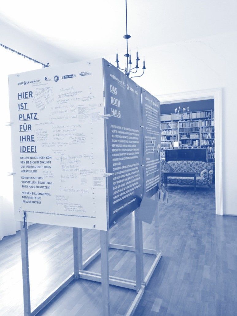 Ein blaues Plakat steht in einem Raum mit Holzboden. Dahinter befindet sich eine Bibliothek mit Regalen voller Bücher und einer Kronleuchter.