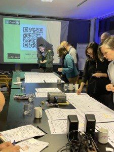 Eine Gruppe von Menschen versammelt sich um einen Tisch und ist in einer Aktivität vertieft. Eine Projektionsleinwand im Hintergrund zeigt einen QR-Code. Papiere, Tassen und ein Beamer stehen auf dem Tisch.