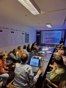 Eine Gruppe von Menschen sitzt in einem Konferenzraum um einen Tisch herum. An der Wand befindet sich eine Projektionsleinwand, die eine Präsentation zeigt. Alle schauen auf den Bildschirm.