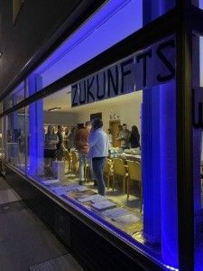 Ein Schaufenster zeigt ein Banner mit der Aufschrift 'ZUKUNFTS'. Drinnen sind Menschen versammelt, einige stehen in der Nähe von Tischen, andere unterhalten sich. Der Raum ist von blauen Lichtern beleuchtet, und der Boden reflektiert die Lichter.