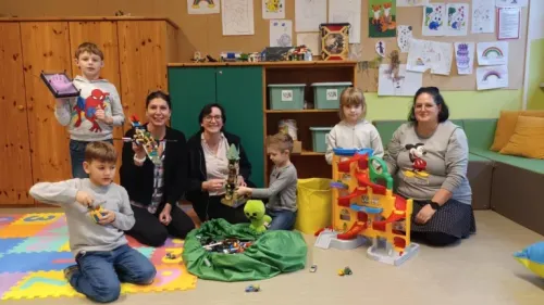 Eine Gruppe von Kindern und Erwachsenen in einem Spielzimmer mit Spielzeug, Aufbewahrungsboxen und Zeichnungen an der Wand.