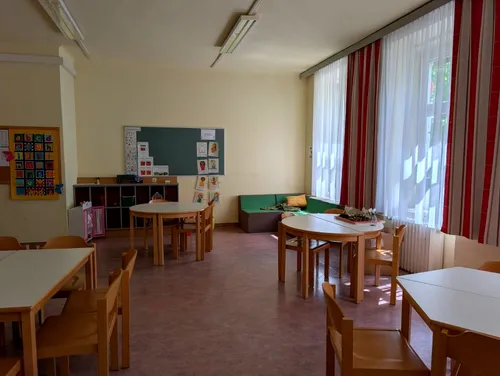 Ein leerer Klassenraum mit Holztischen, Stühlen und einer Tafel mit Bildern. Ein grünes Sofa mit Kissen steht gegen ein Fenster mit weißen Vorhängen.