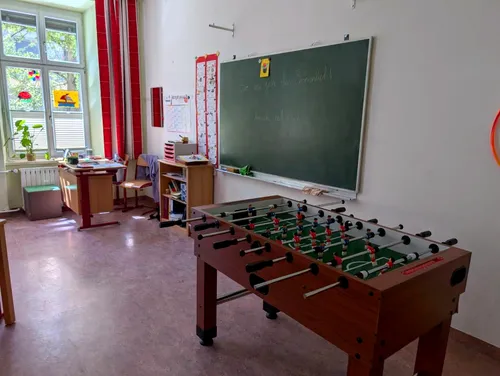 Ein Klassenzimmer hat einen Tischfußball, Schreibtische, einen Schrank und eine Ankündigungstafel an der Wand. Die Tafel hat Schrift darauf.
