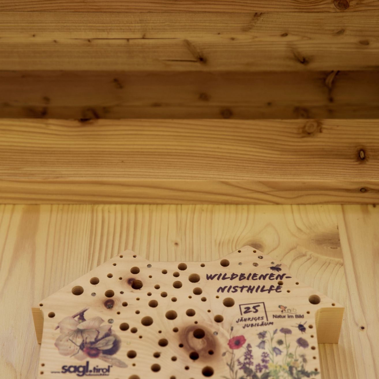 Ein Holzschild mit Löchern, Blumen und Text liegt auf einer Holzoberfläche. Es steht 'WILDBIENEN-NISTHILFE' und '25 JAHRIGES JUBILAUM'. Der Text 'NATUR IM BILD' ist ebenfalls sichtbar.