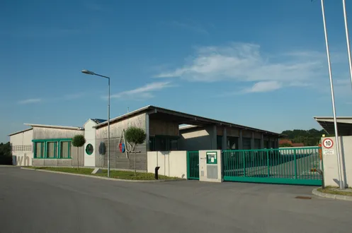 Ein modernes Gebäude mit einem grünen Zaun und Bäumen, unter einem blauen Himmel mit einigen Wolken, mit einer Straßenlaterne auf der linken Seite.