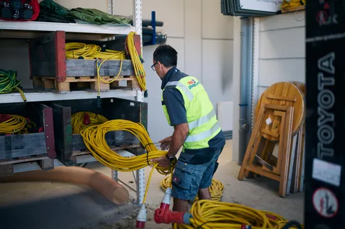 Ein Mann in einer reflektierenden Sicherheitsweste handhabt gelbe elektrische Kabel in einem Lagerbereich, umgeben von Holzpaletten und Regalen.