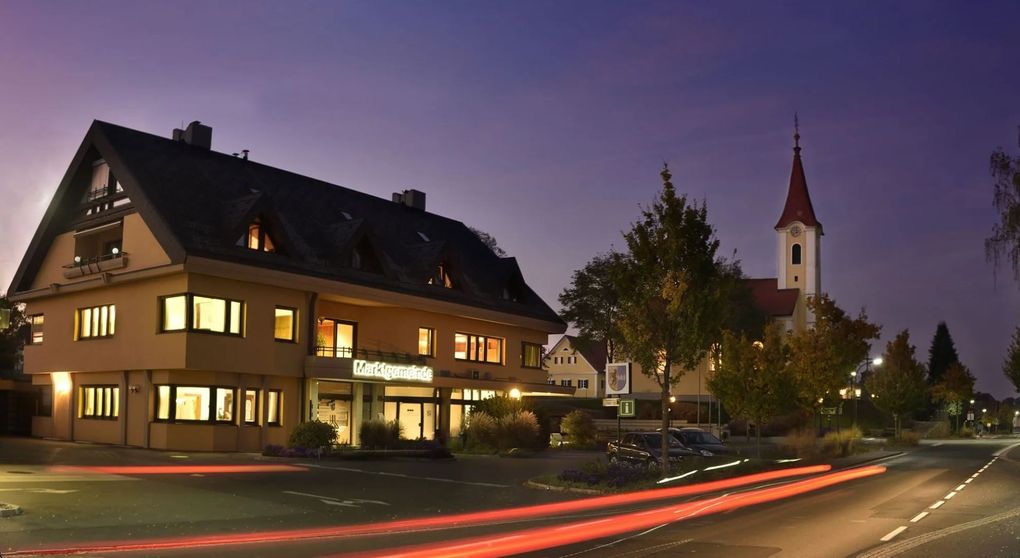  Abschnitt Bild: Die Vorderseite eines Gebäudes ist bei Nacht mit eingeschalteten Lichtern beleuchtet, und ein Kirchturm ist im Hintergrund sichtbar.