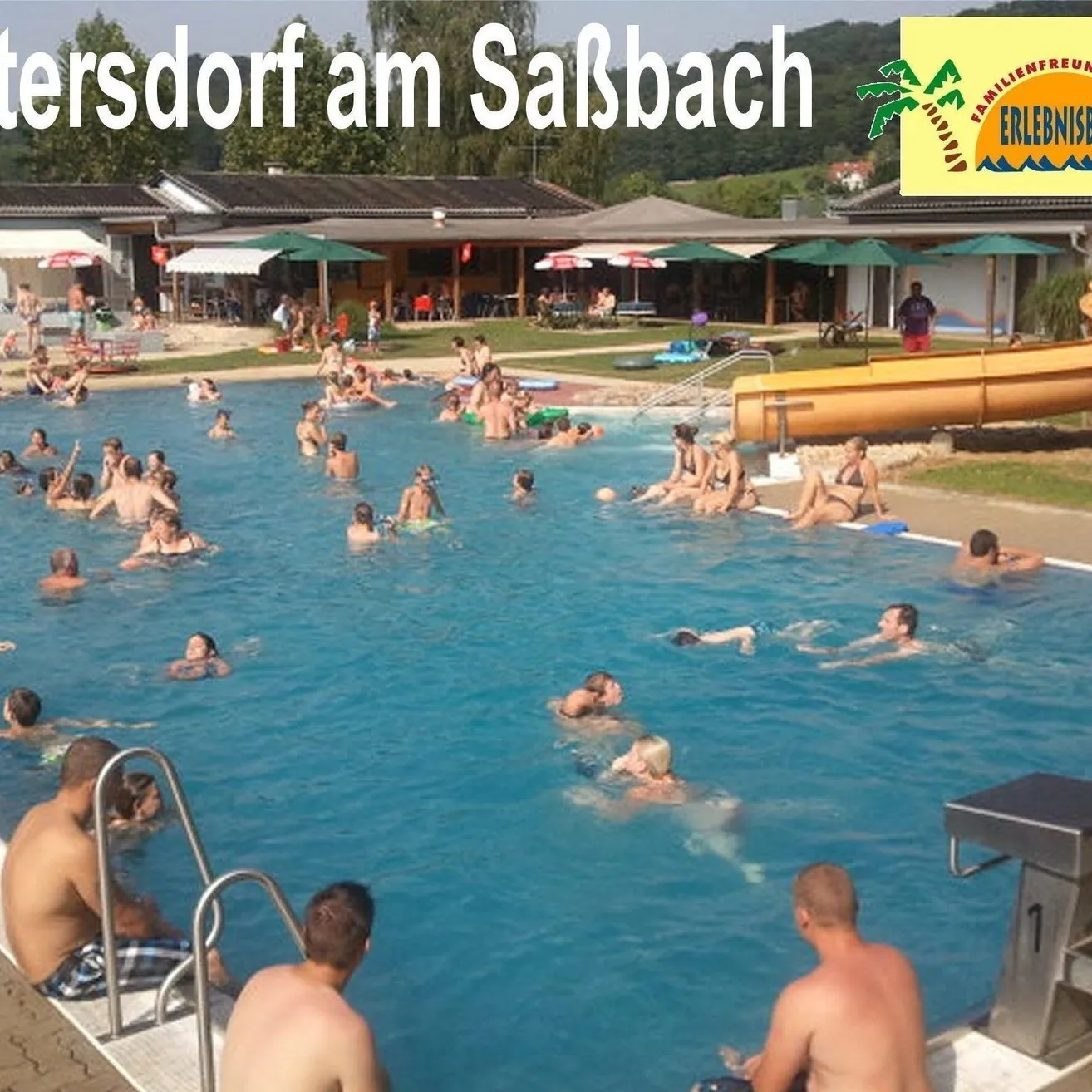 Ein großer Swimmingpool mit Schwimmenden und einer Wasserrutsche im Hintergrund. Der Text lautet 'Mettersdorf am Saisbach' und ein Logo.