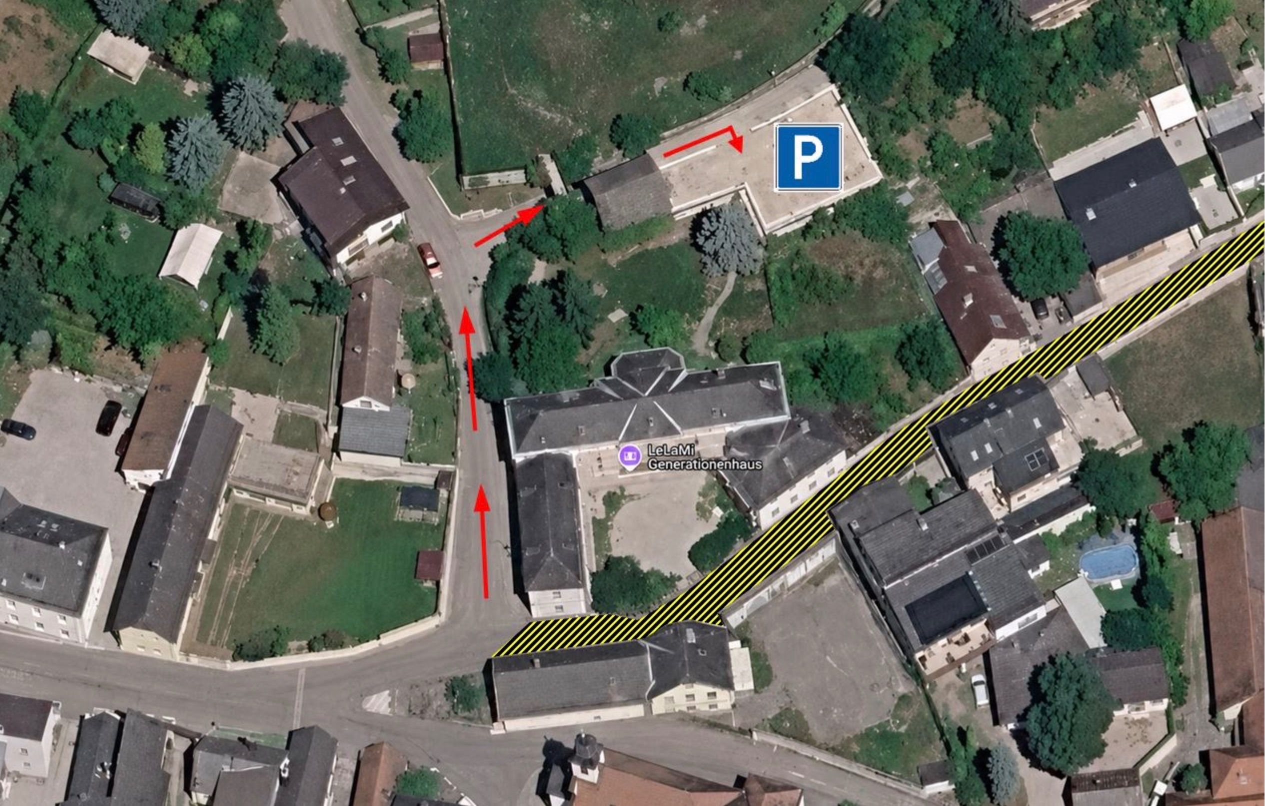 Die Luftaufnahme zeigt ein Gebäude mit einem ausgewiesenen Parkplatz 'P'. Rote Pfeile weisen auf die Eingänge hin. Das Gebäude ist von Häusern und Bäumen umgeben.